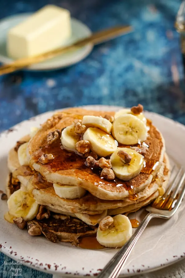 A stack of banana pancakes topped with sliced bananas, toasted walnuts, and maple syrup