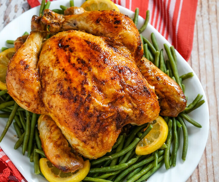 A roasted chicken rests on a bed of green beans with lemons on a white platter.