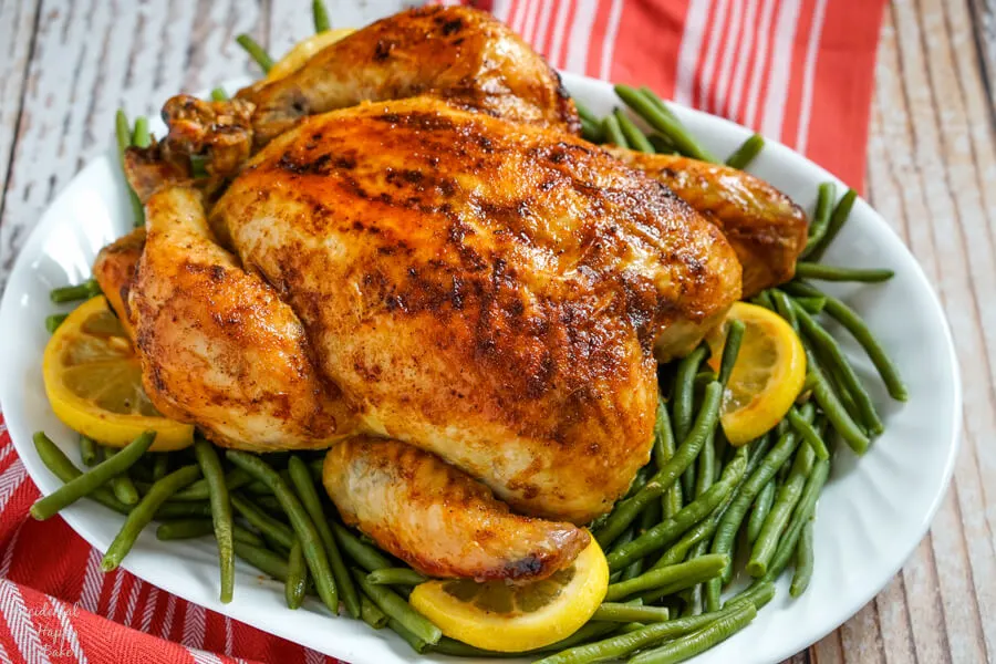 A roasted chicken rests on a bed of green beans with lemons on a white platter.