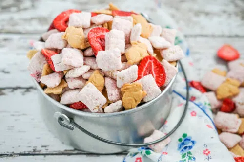 A silver bucket full of strawberry cheesecake muddy buddies with a blue flowered napkin beside it.