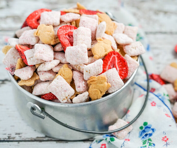 A silver bucket full of strawberry cheesecake muddy buddies with a blue flowered napkin beside it.