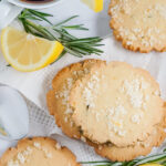 Lemon Rosemary cookies, lemon slices and rosemary sprigs are spread on a white cloth napkin beside a cup of tea.