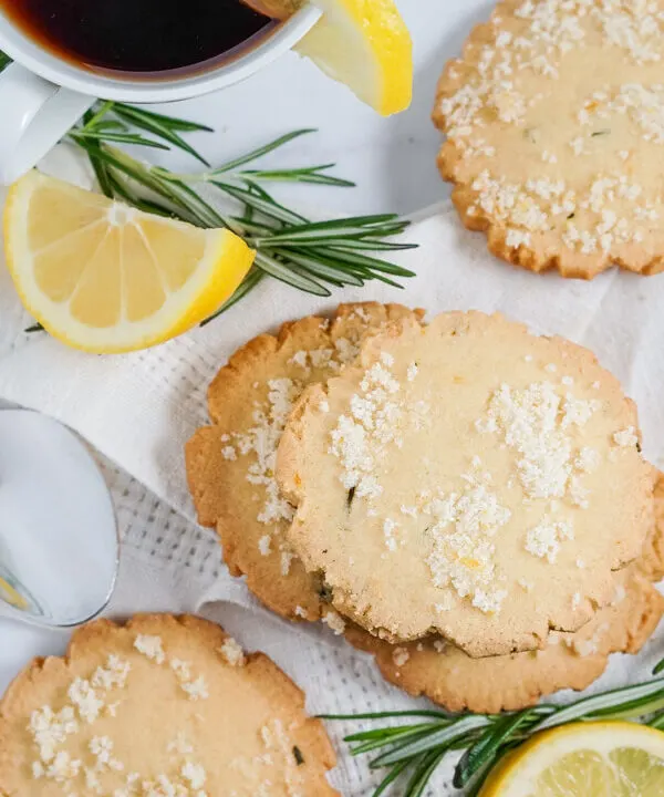 Lemon Rosemary cookies, lemon slices and rosemary sprigs are spread on a white cloth napkin beside a cup of tea.