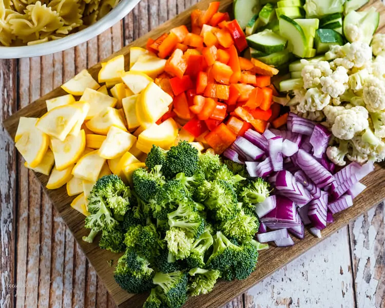 A wooden cutting board loaded with chopped yellow squash, red bell pepper, cucumber, broccoli, red onion, and cauliflower. 