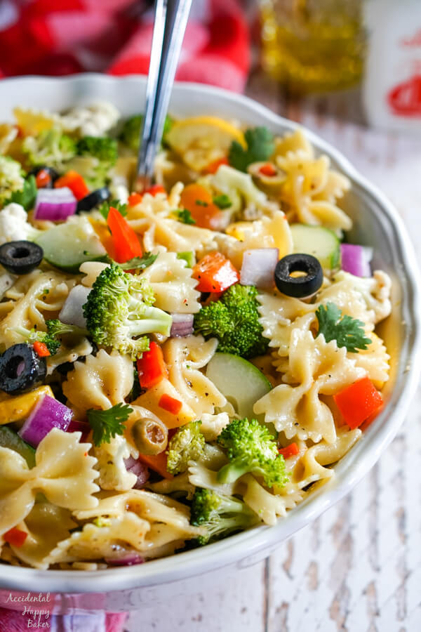 A vertical photo of a white bowl with summer vegetable pasta salad with a serving spoon.