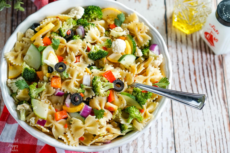 Summer vegetables and farfelle pasta tossed with an Italian dressing in a white bowl. A bottle of olive oil and pepper sit to the side. 