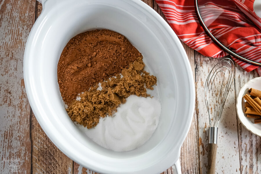 A white slow cooker with cocoa powder, brown sugar and granulated sugar next to a whisk. 