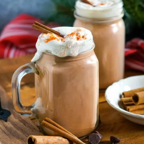 Clear glass mugs of Mexican Hot Cocoa topped with whipped cream and cinnamon sticks on a wooden tray.