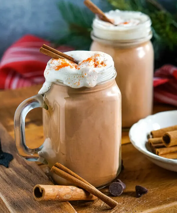 Clear glass mugs of Mexican Hot Cocoa topped with whipped cream and cinnamon sticks on a wooden tray.