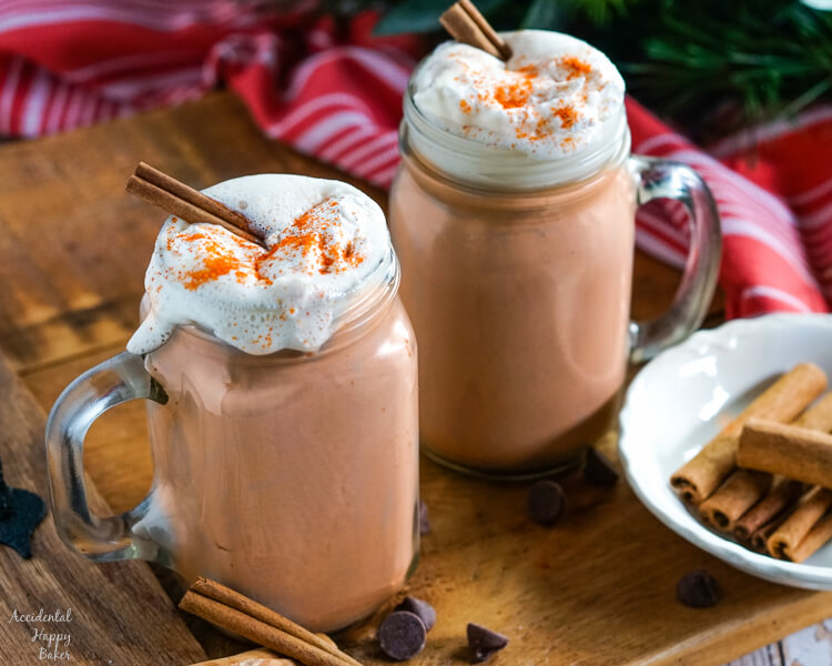 Two mugs of hot cocoa topped with whipped cream, cinnamon sticks, and cayenne pepper. 