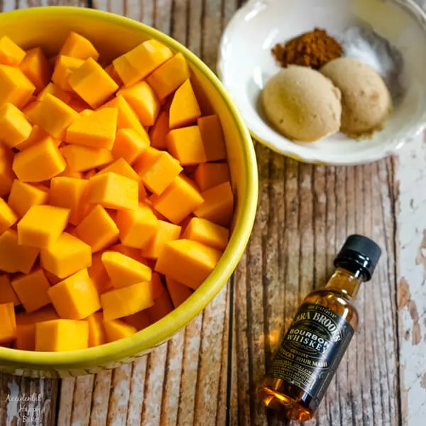 A bowl of peeled and chopped butternut squash sits next to a small bottle of bourbon and a bowl of spices. 