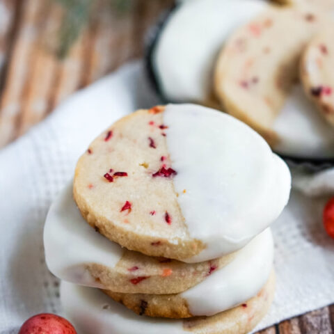 A stack of White Chocolate Cranberry Shortbread Cookies sitting on a white napkin with a cranberry to one side.