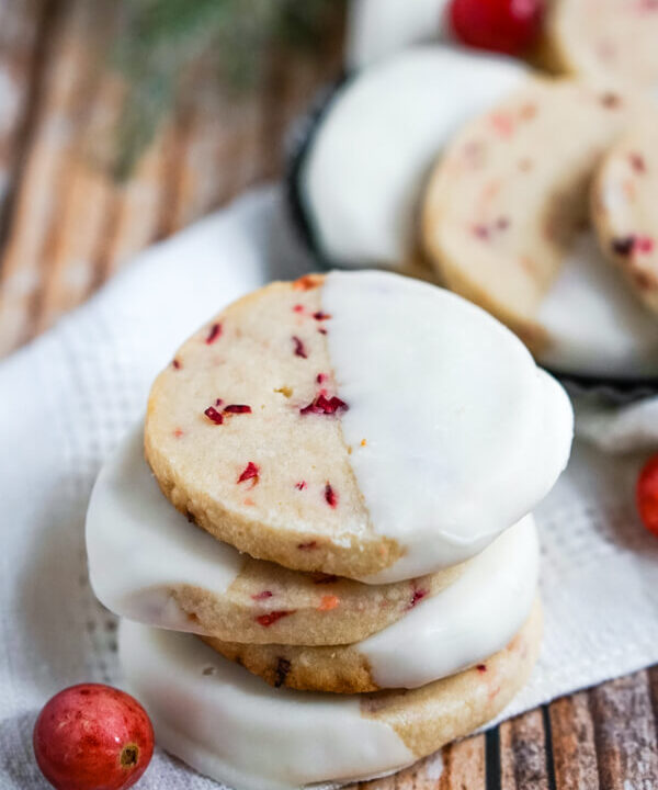 A stack of White Chocolate Cranberry Shortbread Cookies sitting on a white napkin with a cranberry to one side.