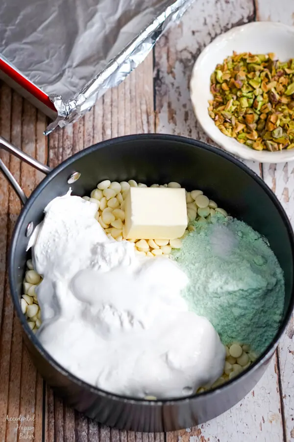 A sauce pan with the ingredients needed to make pistachio swirl fudge sits next to a bowl of chopped pistachios. A foil lined 8x8 pan is in the background. 
