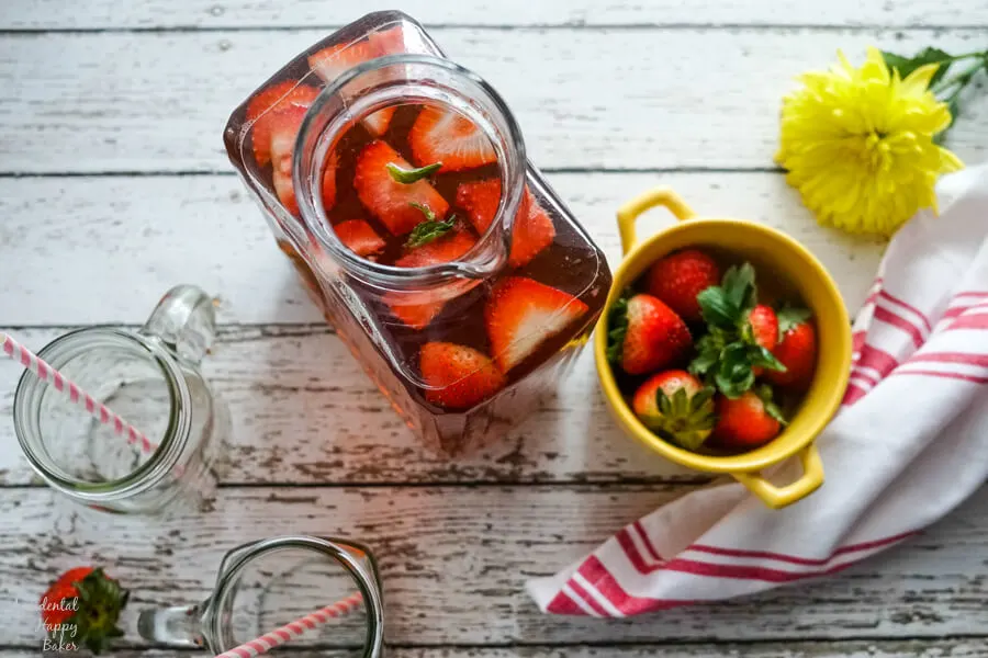 A pitcher of strawberry basil iced tea next to a yellow bowl full of strawberries. 