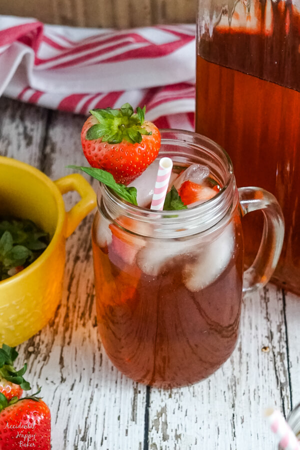 A mason jar cup of strawberry basil iced tea sitting next to a pitcher. 