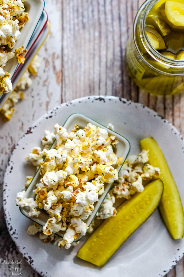 A bowl of popcorn sits on a plate and next to a jar of opened dill pickles. 