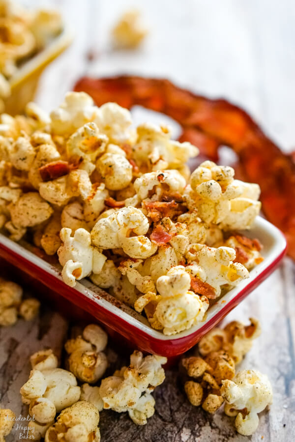 A red bowl full of popcorn with a piece of bacon in the background.