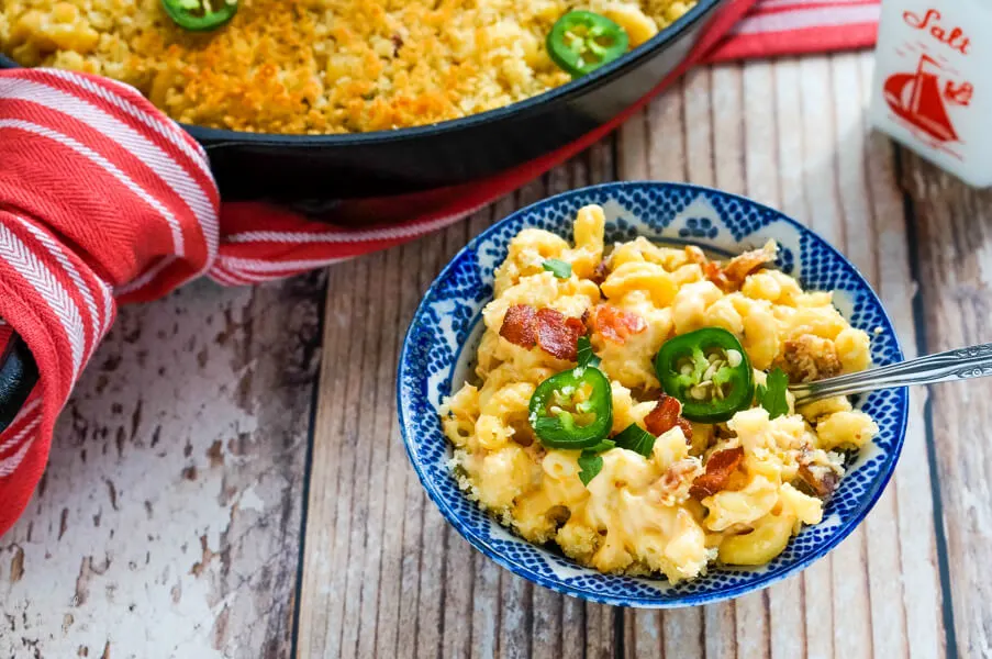 A blue and white bowl full of jalapeno bacon mac and cheese next to a skillet. 