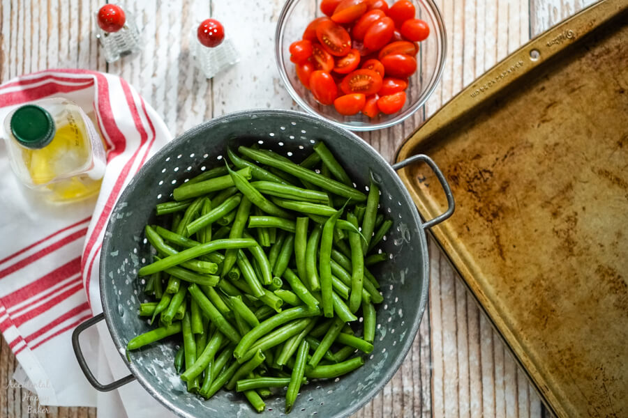 Green beans in a metal colander, next to a bowl of tomatoes, olive oil, salt and pepper.