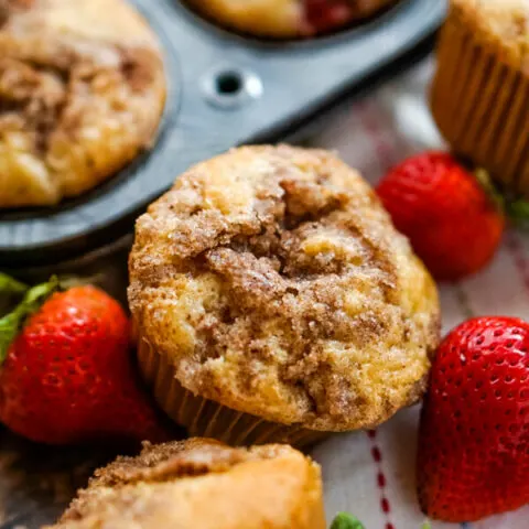 A close up shot of a strawberry rhubarb muffin next to a muffin tin and some fresh strawberries.
