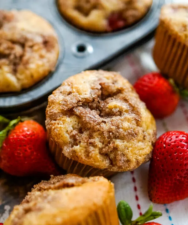 A close up shot of a strawberry rhubarb muffin next to a muffin tin and some fresh strawberries.