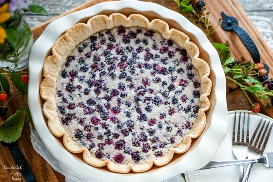 A full blackberry pie in a white pie pan on wooden tray surrounded by blackberries. 