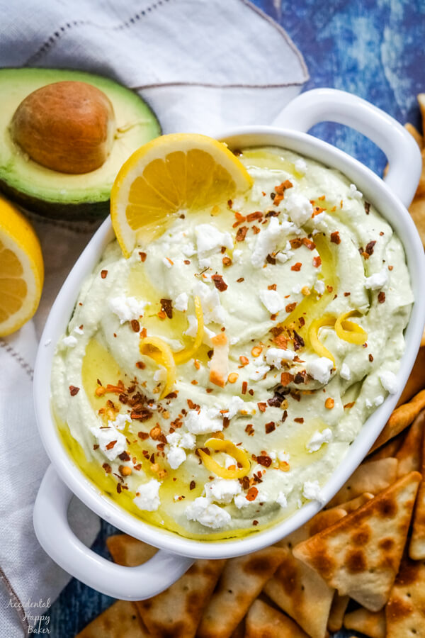 A white bowl full of lemon feta dip surrounded by pita chips and garnished with sliced lemons. 