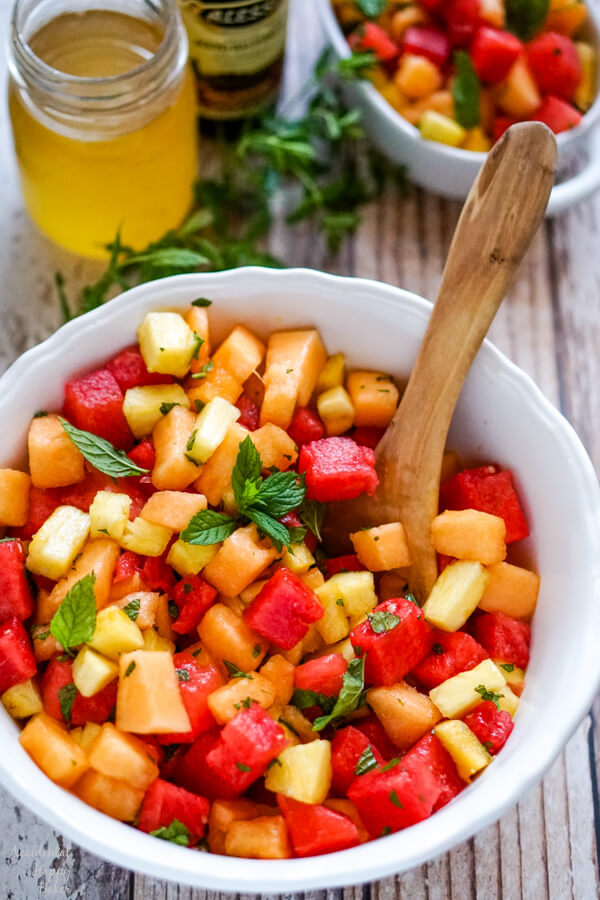 A white bowl of the finished watermelon fruit salad sitting next to a jar of honey and a bottle of balsamic vinegar. 