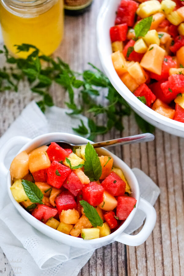 A serving of fruit salad in a small white bowl with several sprigs of fresh mint to the side. 