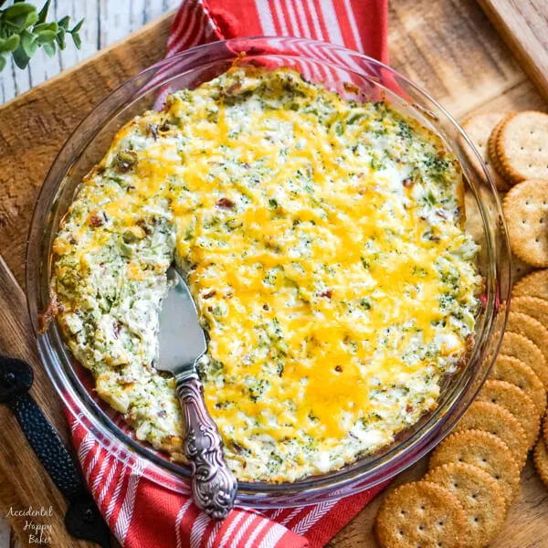 A pie plate of cheesy bacon and broccoli dip hot from the oven on a tray with crackers to one side. 