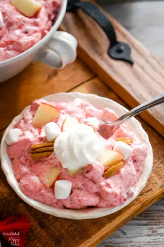 A white serving bowl of Cranberry Apple Fluff Salad topped with a dollop of whipped cream. 