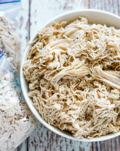 A white bowl full of shredded chicken with 2 freezer bags of chicken next to it. 