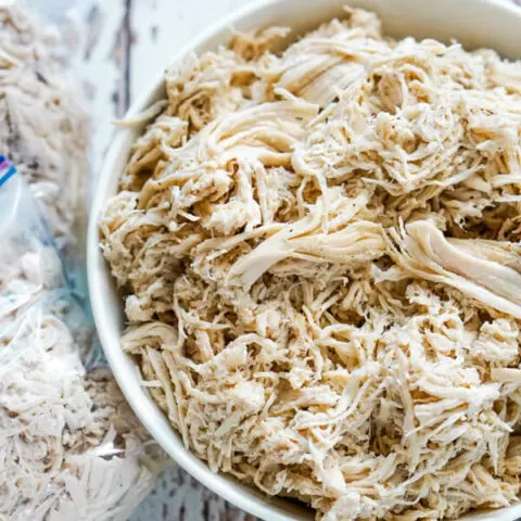 A white bowl full of shredded chicken with 2 freezer bags of chicken next to it.