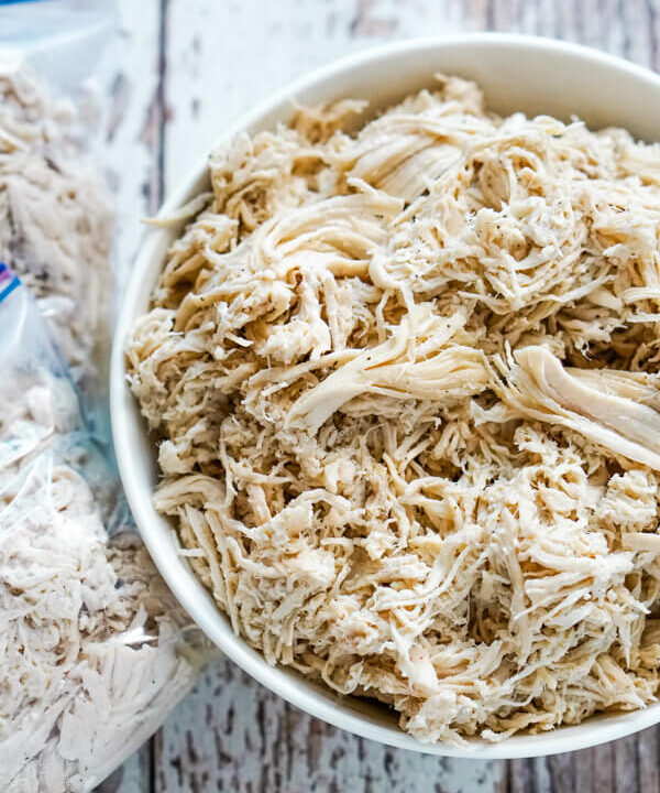 A white bowl full of shredded chicken with 2 freezer bags of chicken next to it.