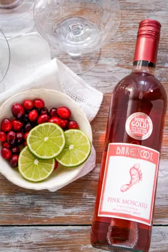 A bottle of pink moscato wine next to a bowl of cranberries and sliced limes. 