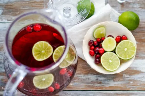 A pitcher of punch with a bowl of sliced limes and cranberries next to it. 