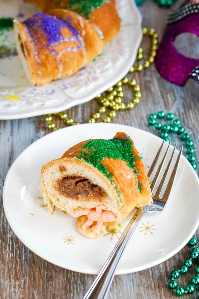 A slice of king cake on a white plate with mardi gras beads in the background. 