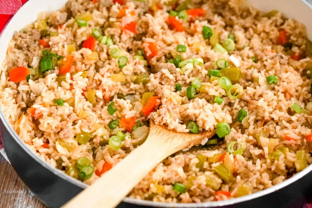 A skillet full of dirty rice with sausage with a wooden spoon holding a scoop of rice. 