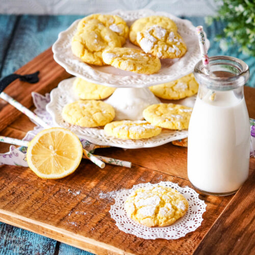 A wooden tray holding a plate of lemon crinkle cookies, a glass milk bottle and a sliced lemon. 