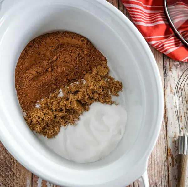 A white slow cooker with cocoa powder, brown sugar, and white sugar inside it.