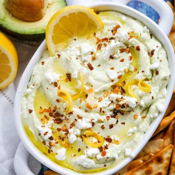 A white bowl full of lemon feta dip surrounded by pita chips and garnished with sliced lemons.