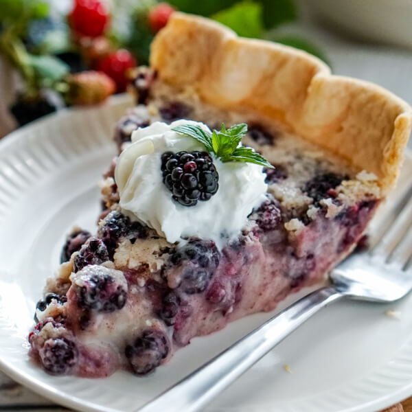 A slice of blackberries and cream pie on a white plate garnished with whipped cream, mint and fresh blackberries.