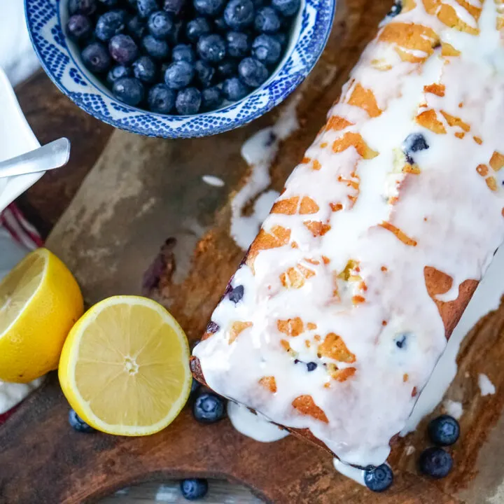 A loaf of lemon blueberry bread with buttermilk glaze on a bread board with lemons and blueberries to one side.