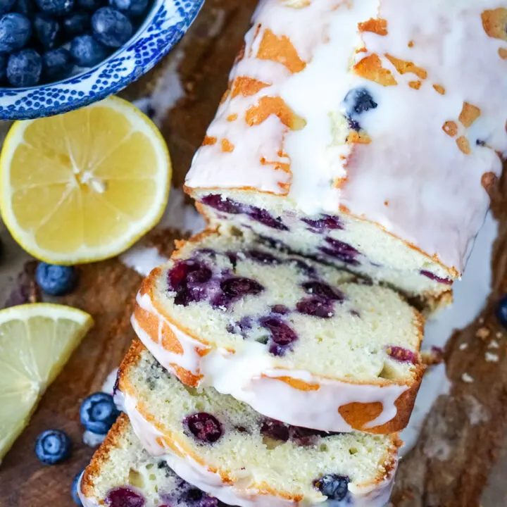 A sliced loaf of lemon blueberry bread with buttermilk glaze on a bread board with a sliced lemon and bowl of blueberries to one side.