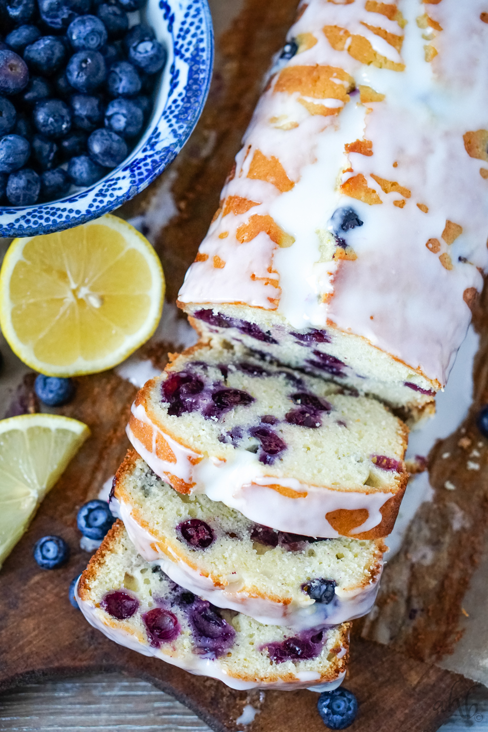 A sliced loaf of lemon blueberry bread with buttermilk glaze over the top on a bread board with sliced lemons and a bowl of blueberries to one side. 