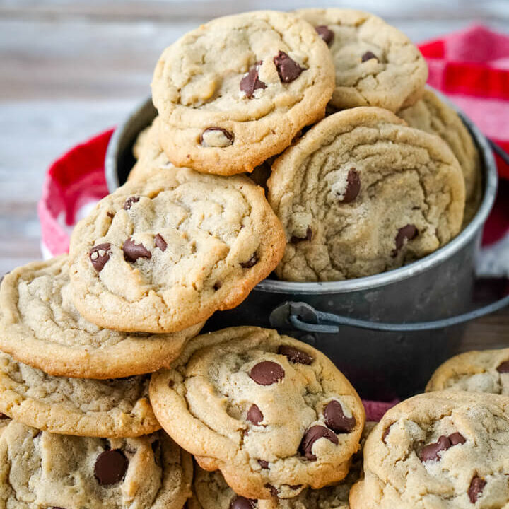 A large stack of chocolate chip cookies next to a metal bucket full of chocolate chip cookies.