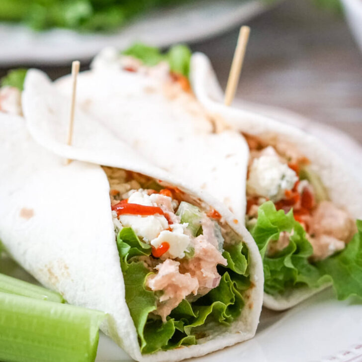 Two Buffalo Tuna Salad wraps with tortillas, lettuce, buffalo tuna salad, blue cheese and extra hot sauce on a white plate and celery sticks to one side.