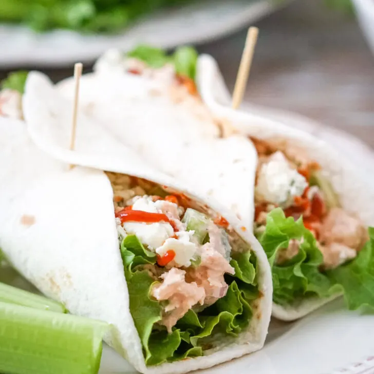 Two Buffalo Tuna Salad wraps with tortillas, lettuce, buffalo tuna salad, blue cheese and extra hot sauce on a white plate and celery sticks to one side.