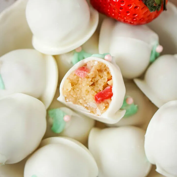 A bowl full of strawberry cheesecake truffles with 2 fresh strawberries to the side.
